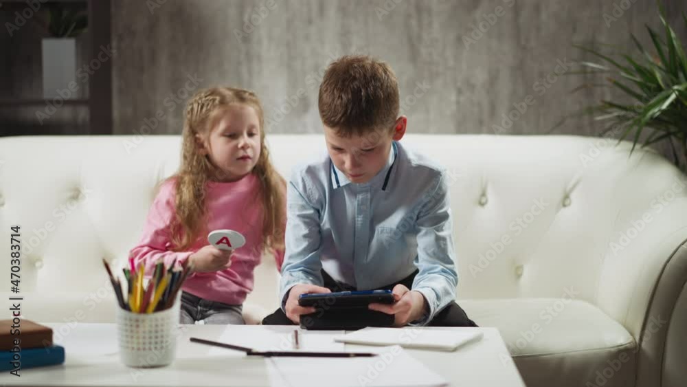 Junior schoolboy with little sister holding paper letters watch online lesson via tablet computer at remote education on sofa in living room slow motion Junior schoolboy with little sister holding paper letters watch online lesson via tablet computer at remote education on sofa in living room slow motion