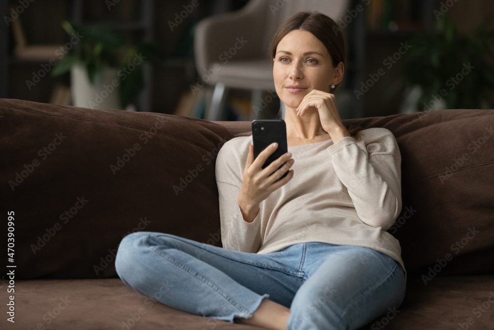 Serene peaceful young woman sit on sofa holds smartphone deep in thoughts looks into distance, thinking out online purchase, distracted from modern tech usage pondering relaxing alone at modern home Serene peaceful young woman sit on sofa holds smartphone deep in thoughts looks into distance, thinking out online purchase, distracted from modern tech usage pondering relaxing alone at modern home