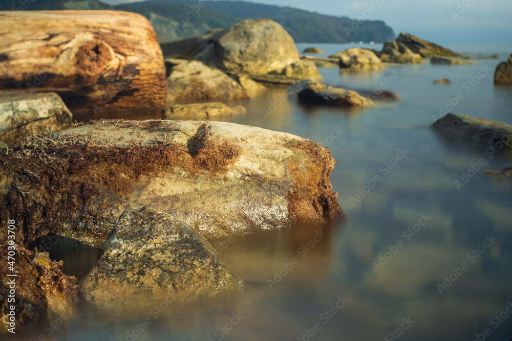 A photo of rocks in seawater with a long exposure. Beautiful reflection on the water. Blue sky and rocky shores in the background.