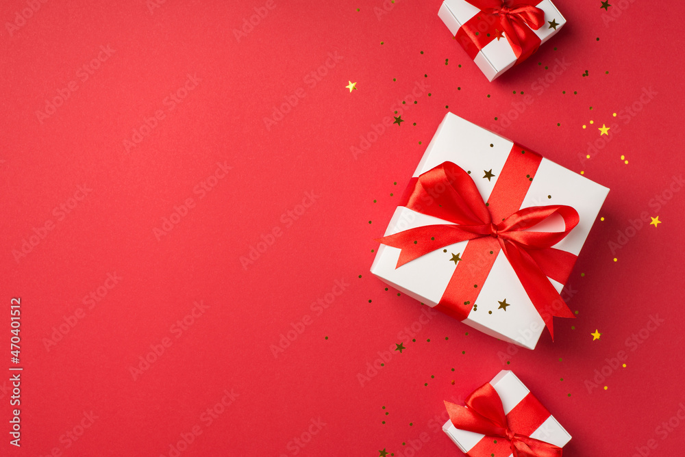 Top view photo of white gift boxes with red silk ribbon bows and golden confetti on isolated red background with blank space