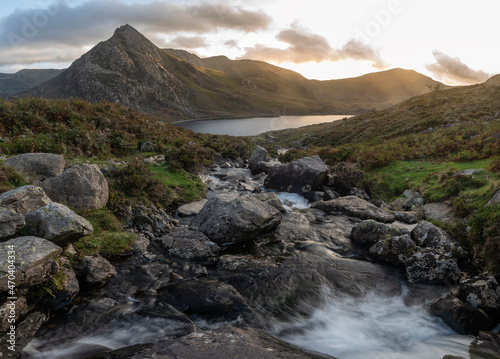 Epic moody Autumn sunset landscape image of Llyn Ogwen and Tryfan in Snowdonia National Park with stream and rocks in foreground
