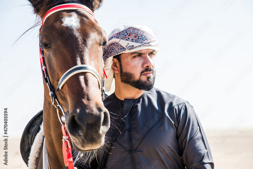 Young adult with Kandura, the emirates traditional clothes, riding his horse in the desert Young adult with Kandura, the emirates traditional clothes, riding his horse in the desert