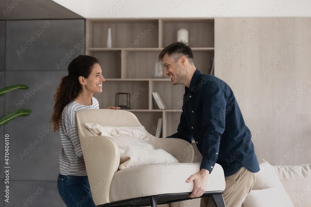 Fototapeta premium Moving day, happy buyers of fashionable furniture, bank mortgage concept. Smiling loving millennial wife and husband carry new armchair into living room, making home design improvements feel satisfied