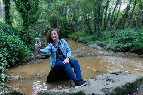 Woman sitting taking a selfie next to a stream running through a gallery forest.