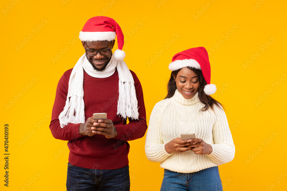 Black Couple Using Smartphones Wearing Santa Hats Over Yellow Background Black Couple Using Smartphones Wearing Santa Hats Over Yellow Background
