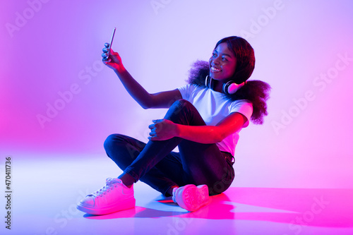 Photography concept. Smiling young black woman taking selfie in neon light, full length photo