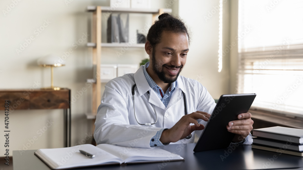 Smiling African American man doctor in white uniform with stethoscope using tablet, sitting at desk in office, physician general practitioner looking at device screen, consulting patient online Smiling African American man doctor in white uniform with stethoscope using tablet, sitting at desk in office, physician general practitioner looking at device screen, consulting patient online