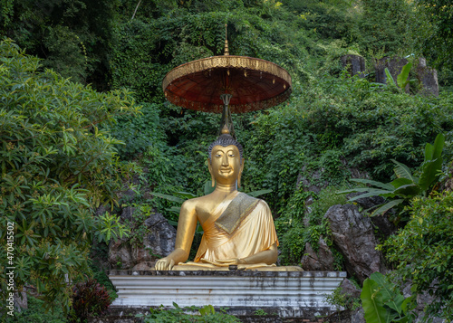 Golden Buddha statue under an umbrella with tropical forest in background on the slopes of Doi Luang mountain, Wat Tham Chiang Dao cave buddhist temple, Chiang Dao, Chiang Mai, Thailand photo
