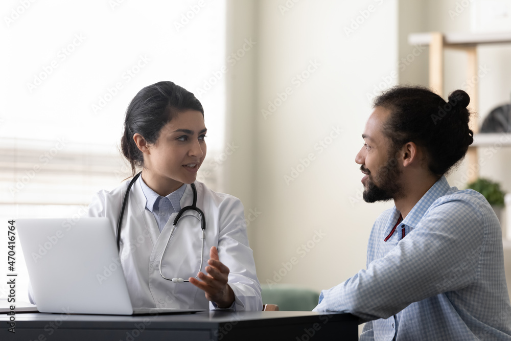 Fototapeta premium Smiling Indian woman doctor consulting African American man patient, therapist practitioner giving recommendation, discussing checkup results or symptoms at meeting, sitting at desk with laptop