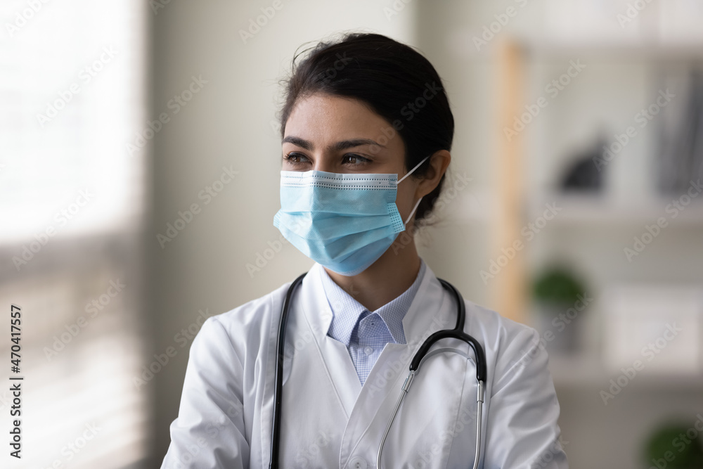 Head shot pensive Indian woman doctor in protective medical mask thinking, looking in distance, thoughtful young female physician practitioner surgeon in white uniform visualizing, planning Head shot pensive Indian woman doctor in protective medical mask thinking, looking in distance, thoughtful young female physician practitioner surgeon in white uniform visualizing, planning