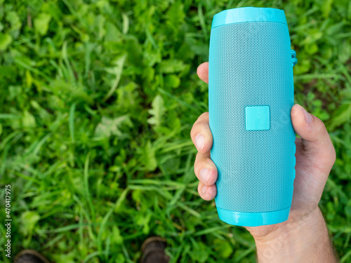 Close-up of a man's hand holding a blue wireless music speaker. There is a thick green grass in the background. Using an outdoor device. Top view, Copy space