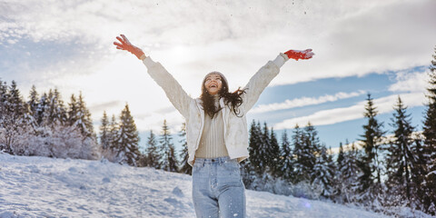 Charming young woman dressed in warm clothes with raised hands in the winter forest outdoors during winter holidays