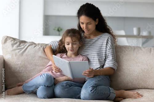 Caring mother with adorable 7s daughter reading book together, sitting on couch at home, loving mom teaching little girl to read, family engaged in educational activity, enjoying fairy tale story