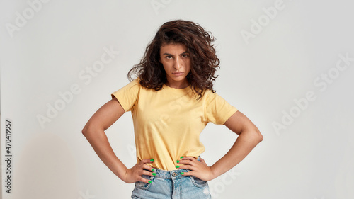 Confident young female model with dark curly hair in casual wear looking at camera while posing isolated over gray background