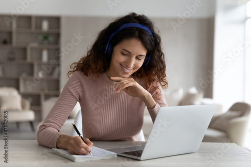 Head shot smiling woman in headphones taking notes watching webinar, studying online at home, happy young female listening to lecture, engaged in internet educational course, remote education