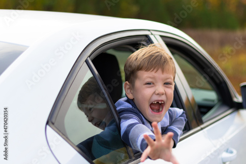 A seven year old cute boy leans out the window of a white car on a warm sunny autumn day against the backdrop of yellow foliage. Selective focus. Portrait