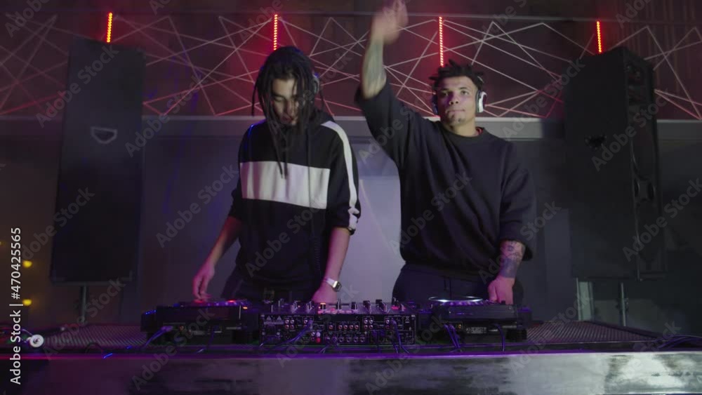 Energetic attractive multiracial male djs in headphones performing live set at stage, mixing music on modern sound mixer and turntables, motivating and cheering up people in nightclub.