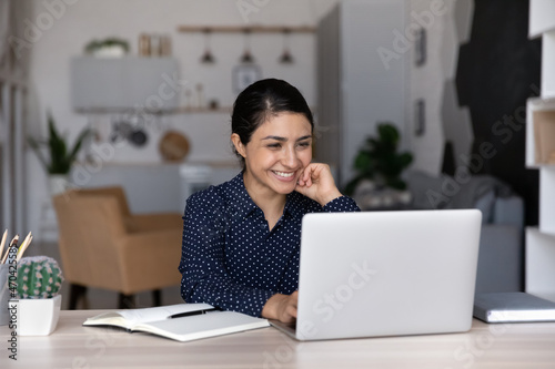 Happy young Indian ethnicity businesswoman looking at computer screen, working distantly at home or office, developing online project, reading email with good news, enjoying studying distantly.