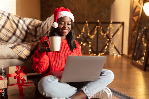 Happy black lady in Santa hat greeting friends with Christmas in video chat on laptop, drinking coffee at home