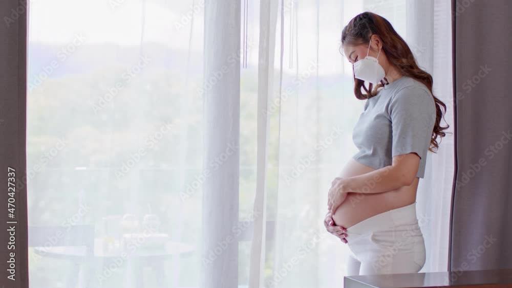 Happy Pregnant Woman wear medical face mask protect virus standing in front of windows stroking big belly with love at cozy home, Pregnancy of young woman enjoy with future life Happy Pregnant Woman wear medical face mask protect virus standing in front of windows stroking big belly with love at cozy home, Pregnancy of young woman enjoy with future life