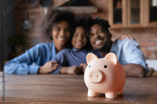 Close up piggybank standing on table with blurred happy African American couple parents and small child daughter on background. Friendly biracial family planning future investment or saving money.