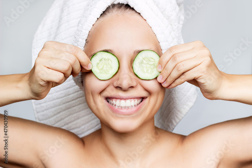 A young happy smiling woman with a white towel on her head after a shower holds cucumber slices covering her eyes making a refreshing face mask isolated on a gray background. Skin care, cosmetology