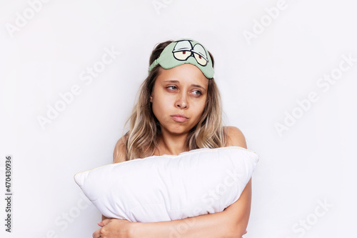 A young dissatisfied sleepless blonde woman with dark circles under the eyes in a green sleep mask with sleepy eyes holds a pillow looking away isolated on a white background. Insomnia