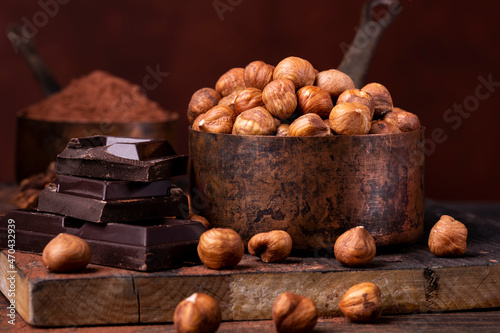 In the foreground, a group of shelled hazelnuts in an aged metal measuring cup, pieces of dark chocolate and cocoa powder on a dark background