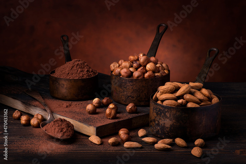 Still life. in the foreground with dark background, vintage metal measuring spoons with shelled hazelnuts and almonds and cocoa powder photo