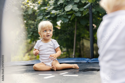 Jumping on a trampoline. A cute toddler boy or a girl in white baby bodysuits is playing in the yard and jumping on the trampoline. Toddlers with blonde hair and blue eyes. Growing up in the home yard
