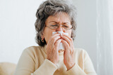 Portrait of senior sick woman sneezing, snotting, coughing into napkin while sitting at home. Flu, runny nose, colds, allergies, virus, seasonal illness in an elderly woman