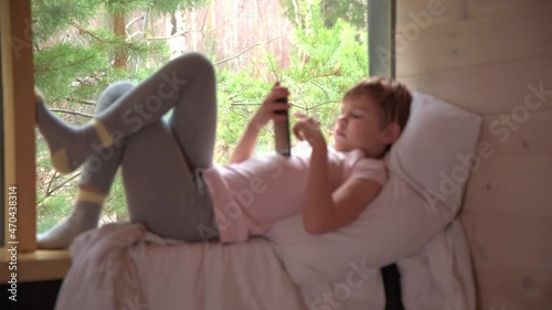 The child looks into a smartphone. The girl settled down comfortably on the windowsill, spread a blanket and a pillow. Outside the window you can see the forest, trees, greenery.