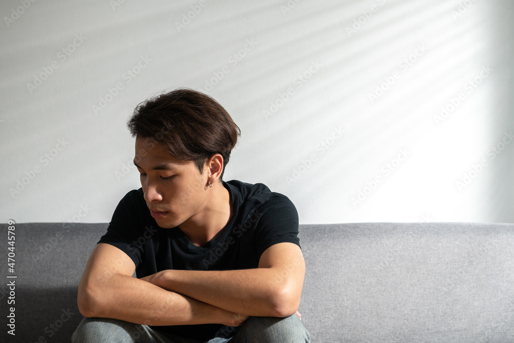 An upset Asian young man sitting alone near a window on a couch at home office sad and looking down, having problems. Copy space. Loneliness, depression, suffering, financial hangover concept ideas