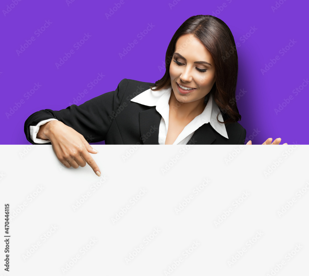 Happy smiling brunette business woman in confident black suit, standing behind, peeping from blank banner or mock up signboard, showing pointing copy space, over violet purple color background. Happy smiling brunette business woman in confident black suit, standing behind, peeping from blank banner or mock up signboard, showing pointing copy space, over violet purple color background.