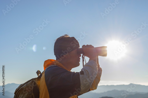 Young male with backpack looking through the binoculars on mountain at beautiful sunset background.