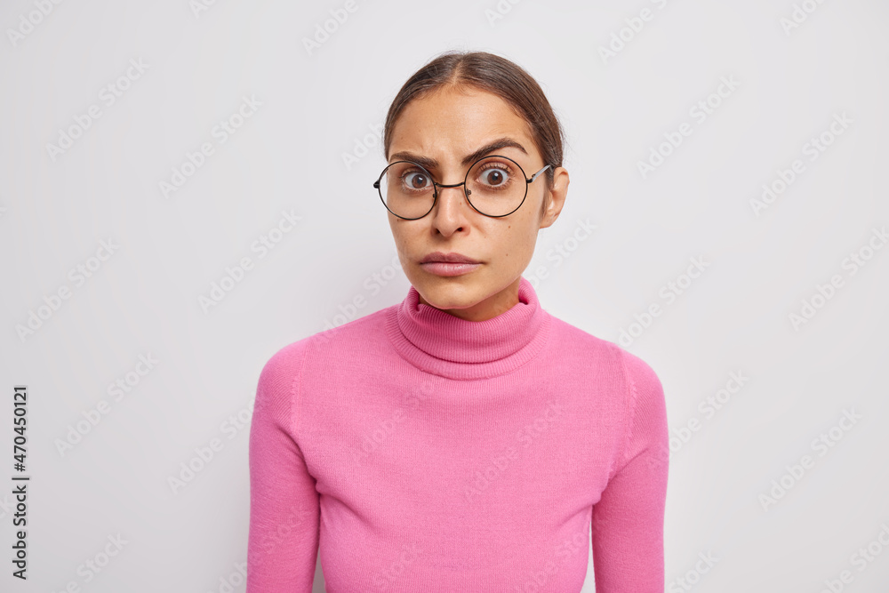 Portrait of serious woman looks attentively at camera hears something surprising wears round spectacles and pink poloneck isolated over white background reacts on bad news. Face expressions concept