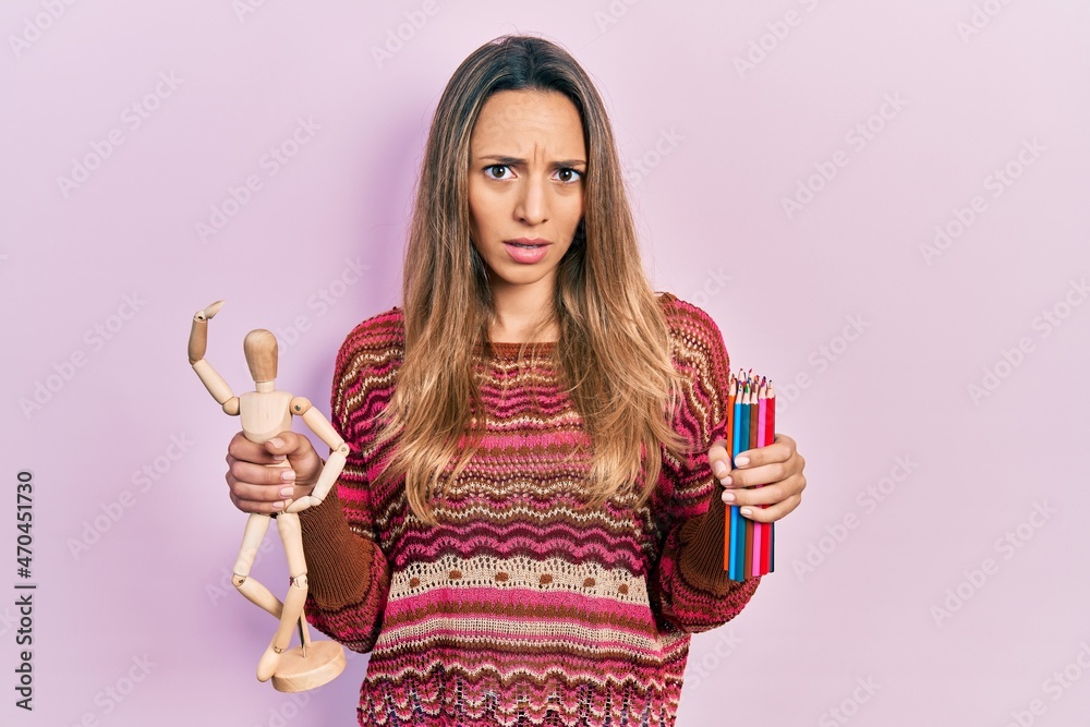 Beautiful hispanic woman holding small wooden manikin and colored pencils in shock face, looking skeptical and sarcastic, surprised with open mouth Beautiful hispanic woman holding small wooden manikin and colored pencils in shock face, looking skeptical and sarcastic, surprised with open mouth