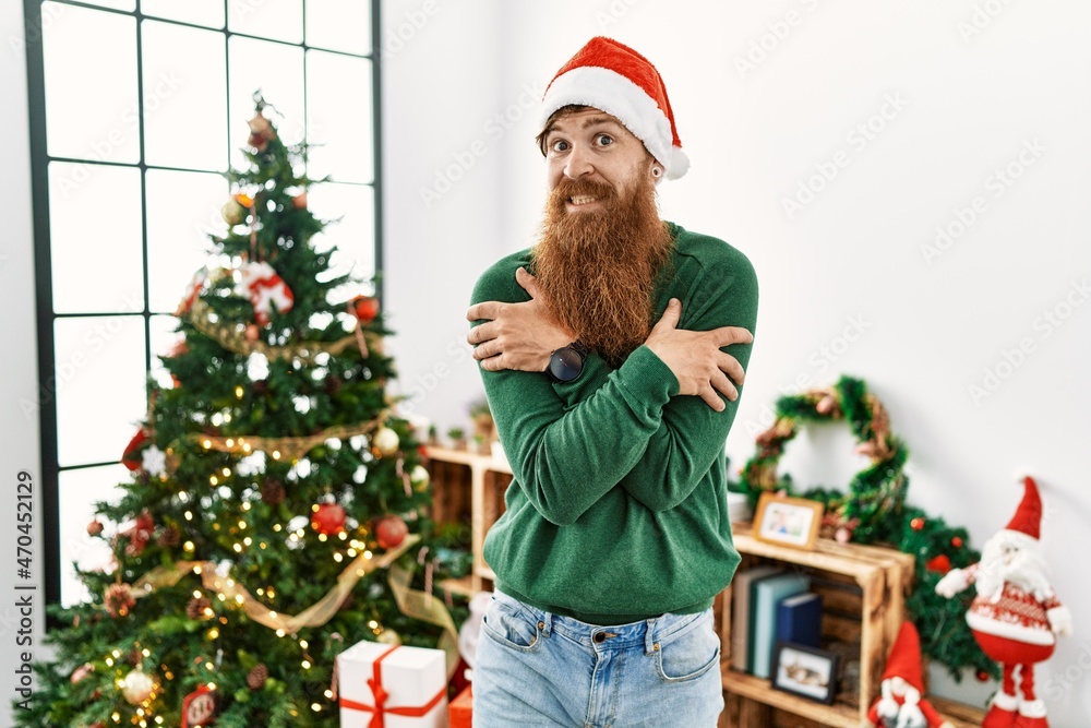 Redhead man with long beard wearing christmas hat by christmas tree shaking and freezing for winter cold with sad and shock expression on face Redhead man with long beard wearing christmas hat by christmas tree shaking and freezing for winter cold with sad and shock expression on face