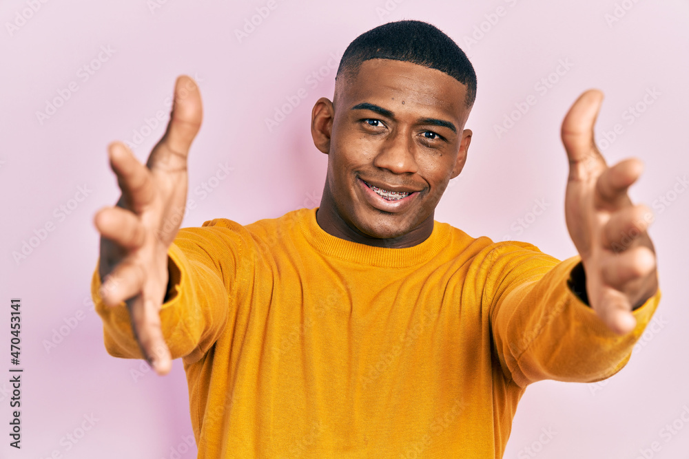 Young black man wearing casual yellow sweater looking at the camera smiling with open arms for hug. cheerful expression embracing happiness. Young black man wearing casual yellow sweater looking at the camera smiling with open arms for hug. cheerful expression embracing happiness.