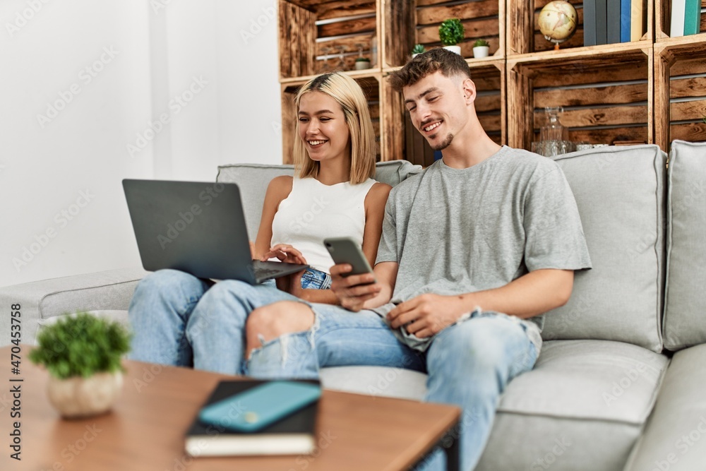Young caucasian couple smiling happy using laptop and smartphone sitting on the sofa at home. Young caucasian couple smiling happy using laptop and smartphone sitting on the sofa at home.
