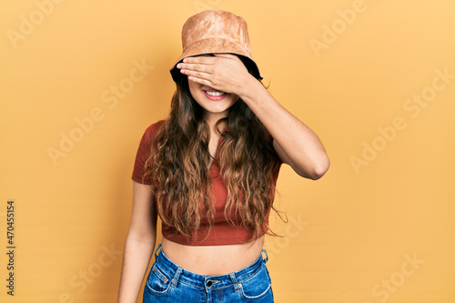 Young hispanic girl wearing casual clothes and hat smiling and laughing with hand on face covering eyes for surprise. blind concept.
