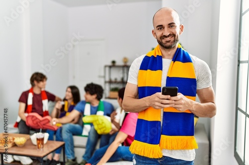 Group of young hispanic friends watching and supporting soccer match. Man smiling happy and using smartphone at home.
