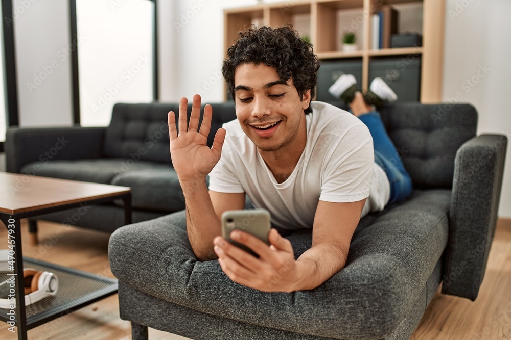 Young hispanic man having video call using smartphone lying on the sofa at home. Young hispanic man having video call using smartphone lying on the sofa at home.