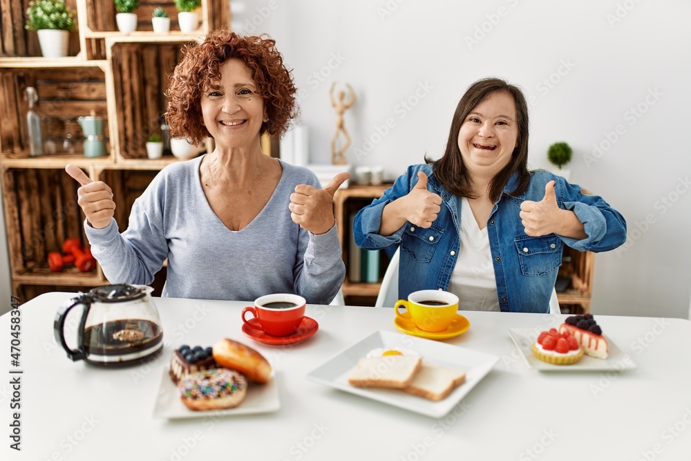 Family of mother and down syndrome daughter sitting at home eating breakfast success sign doing positive gesture with hand, thumbs up smiling and happy. cheerful expression and winner gesture.