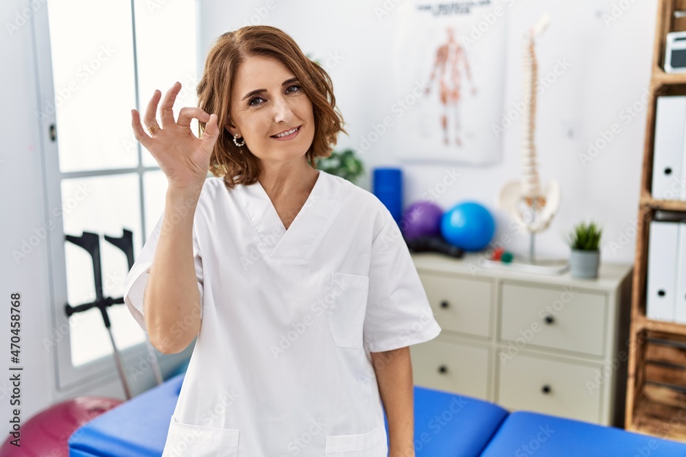 Middle age physiotherapist woman working at pain recovery clinic smiling positive doing ok sign with hand and fingers. successful expression. Middle age physiotherapist woman working at pain recovery clinic smiling positive doing ok sign with hand and fingers. successful expression.
