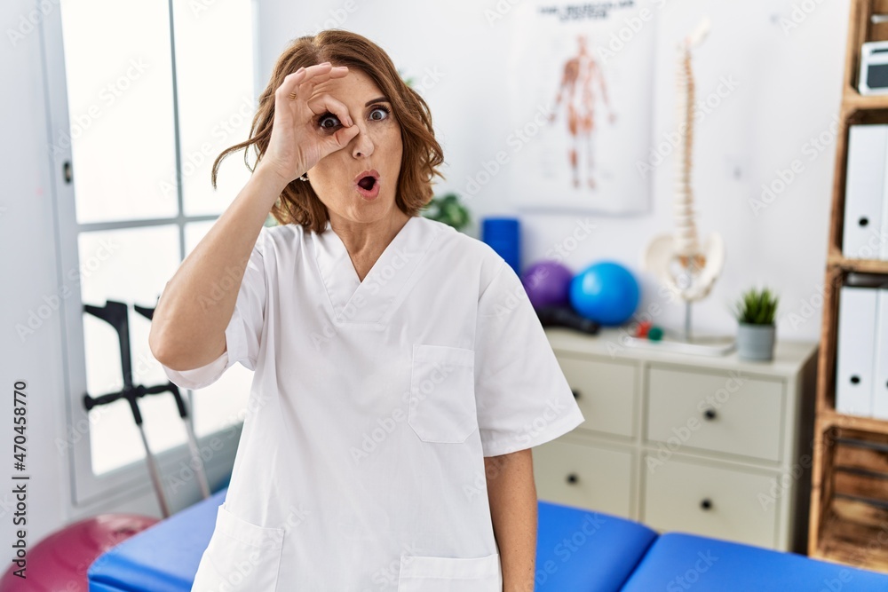 Middle age physiotherapist woman working at pain recovery clinic doing ok gesture shocked with surprised face, eye looking through fingers. unbelieving expression. Middle age physiotherapist woman working at pain recovery clinic doing ok gesture shocked with surprised face, eye looking through fingers. unbelieving expression.
