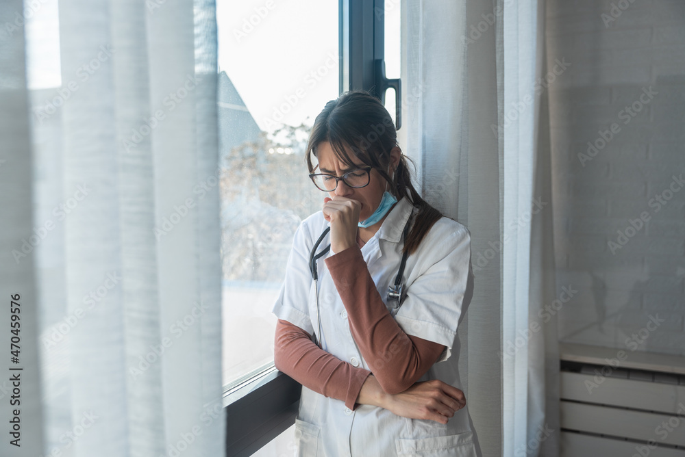 Young sad woman doctor taking a break standing and looking through the window wearing medical protective mask at work feeling tired and depressed after days of overworking shift at the hospital Young sad woman doctor taking a break standing and looking through the window wearing medical protective mask at work feeling tired and depressed after days of overworking shift at the hospital