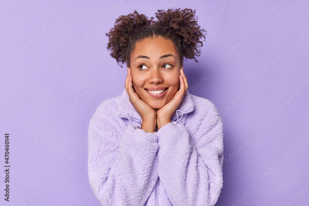 Good looking cheerful young woman with two curly hair buns keeps hands on cheeks smiles pleasantly focused away wears fur winter coat isolated over purple background. People and emotions concept Good looking cheerful young woman with two curly hair buns keeps hands on cheeks smiles pleasantly focused away wears fur winter coat isolated over purple background. People and emotions concept