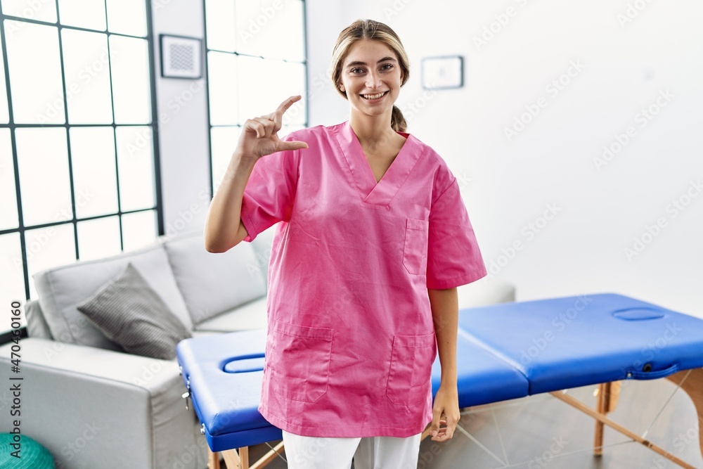 Young blonde woman working as physiotherapist at home smiling and confident gesturing with hand doing small size sign with fingers looking and the camera. measure concept.