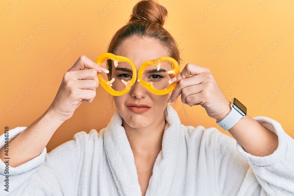Young blonde woman wearing bathrobe holding yellow pepper over eyes skeptic and nervous, frowning upset because of problem. negative person. Young blonde woman wearing bathrobe holding yellow pepper over eyes skeptic and nervous, frowning upset because of problem. negative person.